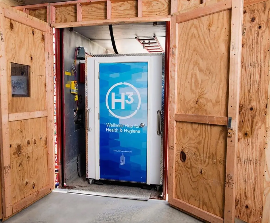 Wooden shelter framing an H³ Wellness Hub unit labeled for Health & Hygiene, with open doors revealing internal electrical components and a blue panel featuring the H³ logo.