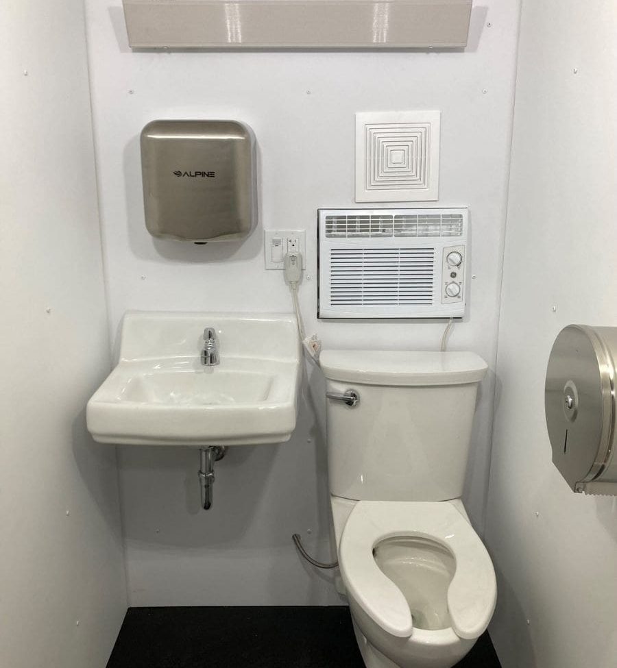 Interior of the H³ Wellness Pod with toilet, toilet paper dispenser, soap dispenser, AC and vent and lighting
